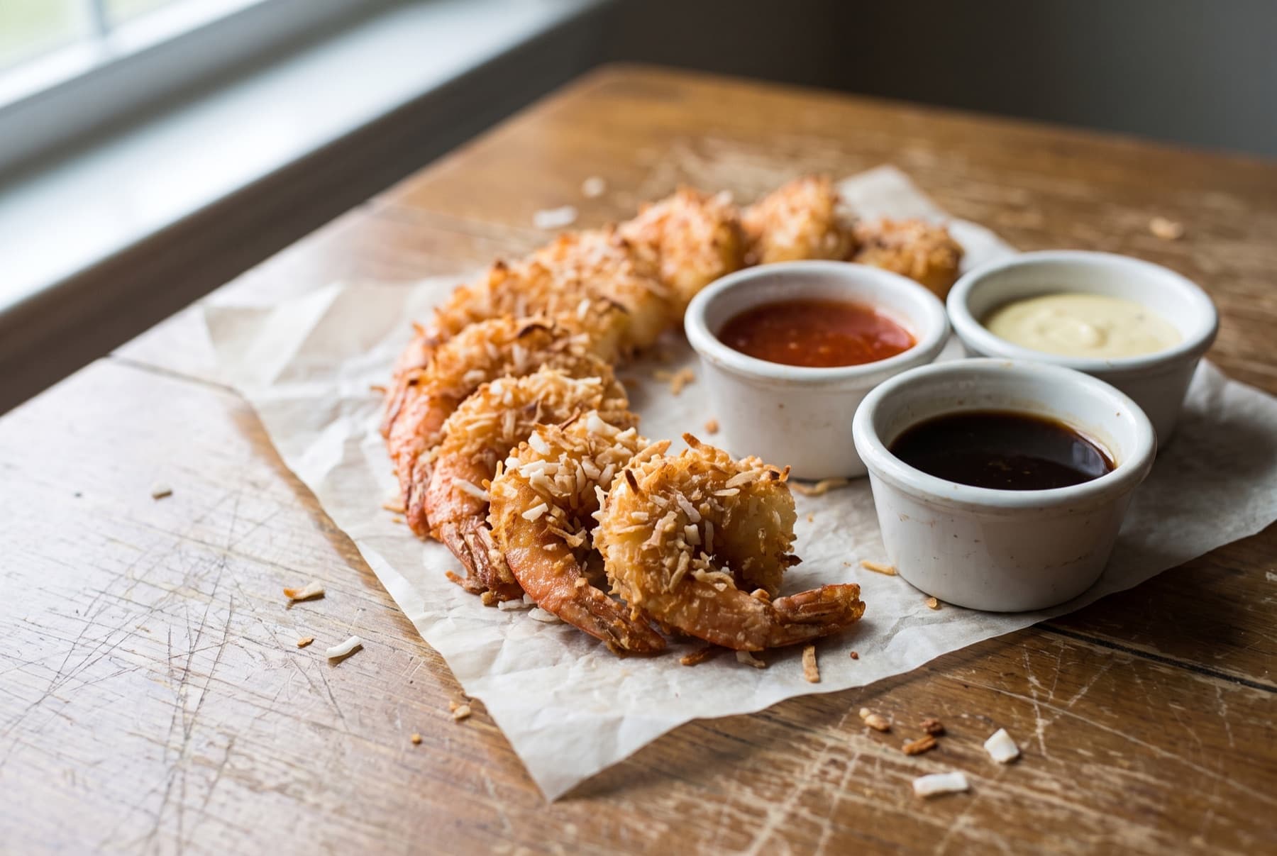 Crispy golden coconut shrimp with visible coconut flakes, fanned on parchment with three small dipping sauces in white ramekins.